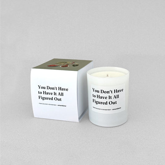 Unundone’s ‘You Don’t Have to Have It All Figured Out’ candle in a glass jar standing next to packaging, photographed on a neutral grey studio background.