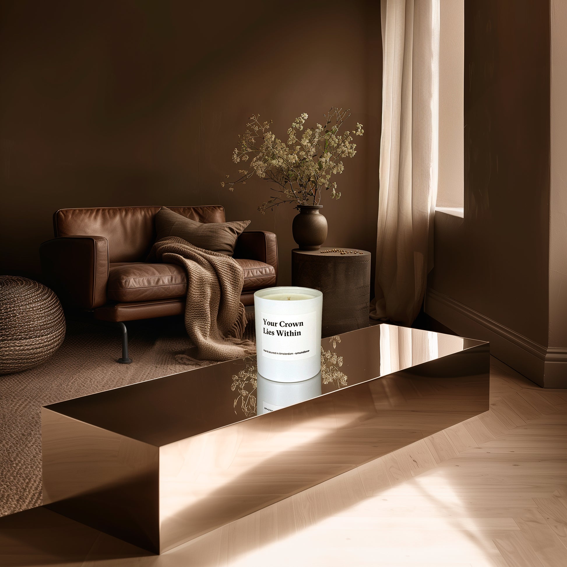 Mood image of Unundone’s ‘Your Crown Lies Within’ candle placed on a reflective podium in a warm-toned room with a leather couch and floral arrangement.